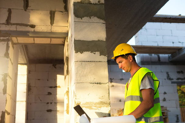 Construction engineer on the construction site of a house made of porous concrete blocks works at a computer in a reflective safety vest and hardhat. Design, construction, drawings project, check - Φωτογραφία, εικόνα