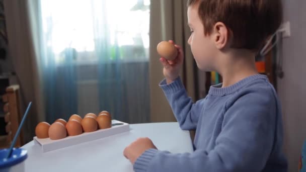 Een klein kind schildert een paaseieren aan tafel op een blauwe achtergrond.Little Boy schildert thuis paaseieren met verf. Creativiteit met Pasen voor kinderen. Een kind versiert paaseieren. - Video