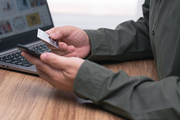 Человек, держащий в руках кредитную карту и смартфон для совершения онлайн-банкинга, совершения оплаты или покупки в интернет-магазине и ноутбуке на столе - Фото, изображение
