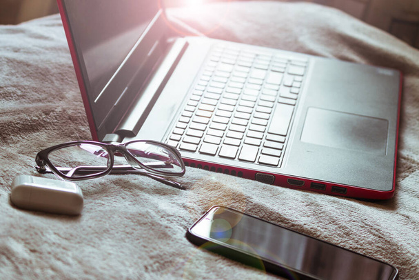 Um laptop, óculos, fones de ouvido e um telefone ficam na cama enquanto trabalham remotamente. - Foto, Imagem