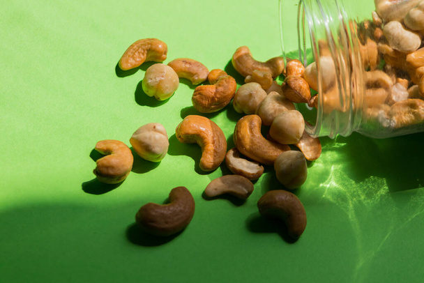 Scattered nuts from a glass jar on a green background. Hazelnuts and cashews. - Valokuva, kuva