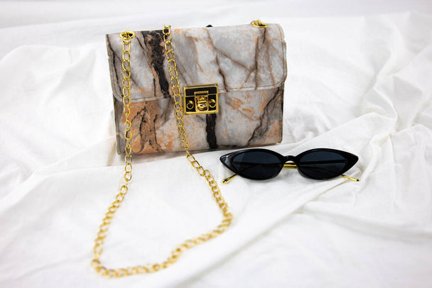 Жіноча сумка з чорними сонцезахисними окулярами на білому тлі. Шкіряна сумка на зібраній тканині. Бічна концепція моди вишуканих модних окулярів та розкішної справжньої шкіряної сумочки
 - Фото, зображення