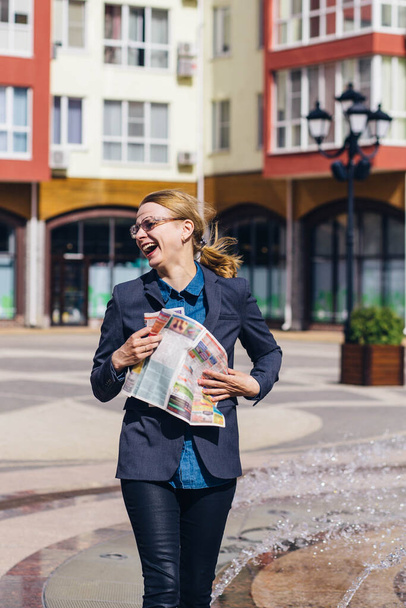 blonde girl in a gray jacket and glasses jumping and having fun with a newspaper in her hands on the street. - Foto, imagen