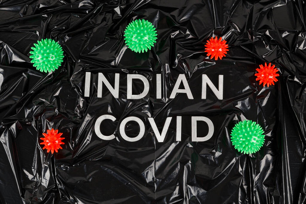 palabras indio covid establecido con letras de metal plateado sobre arrugado negro bolsa de plástico de fondo con pequeños modelos de virus - Foto, Imagen