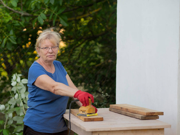 Senior caucasian woman in a blue t-shirt and red gloves is sanding a board with a sander on a layout in a garden near a white wall at home - Photo, Image
