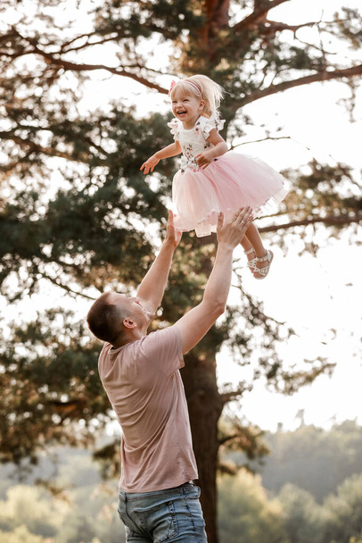 Father throws baby in nature, girl flies in the sky. Portrait dad with child together. Daddy, little daughter outdoors. Young father with baby girl walk in park. Family holiday in garden or forest - Foto, afbeelding