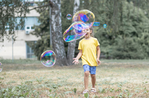 Szczęśliwa dziewczyna łapie duże bańki mydlane, dziecko bawi się na ulicy, lekkie balony unoszą się w powietrzu. - Zdjęcie, obraz