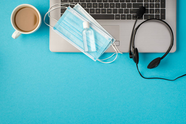 Bovenaanzicht foto van twee medische gezichtsmaskers en transparante reiniger fles op laptop hoofdtelefoon en kopje drank op geïsoleerde pastelblauwe achtergrond met copyspace - Foto, afbeelding