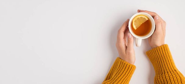 First person top view photo of female hands in yellow sweater touching white cup of tea with lemon slice on isolated white background with copyspace - 写真・画像
