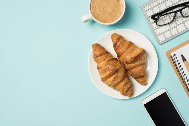 Top view photo of workplace keyboard glasses notebook pen mobile phone cup of frothy coffee and plate with two croissants on isolated pastel blue background with copyspace - Fotoğraf, Görsel