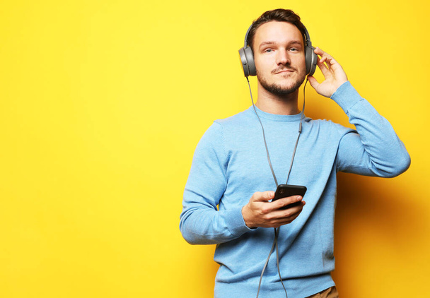 Young handsome man wearing blue sweater listening to music in headphones, yellow studio background, isolated, smiling, happy. - Фото, изображение