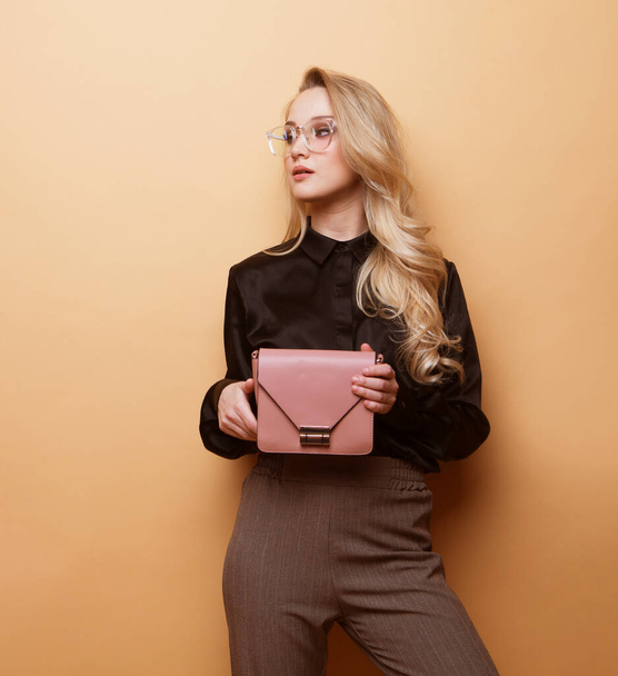Beautiful blonde girl in a black blouse and brown trousers holds a fashionable pink handbag over beige background - 写真・画像