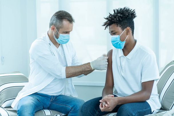 Lekarz w masce trzyma strzykawkę ze szczepionką COVID-19 Afroamerykaninowi w stacji szczepień..  - Zdjęcie, obraz