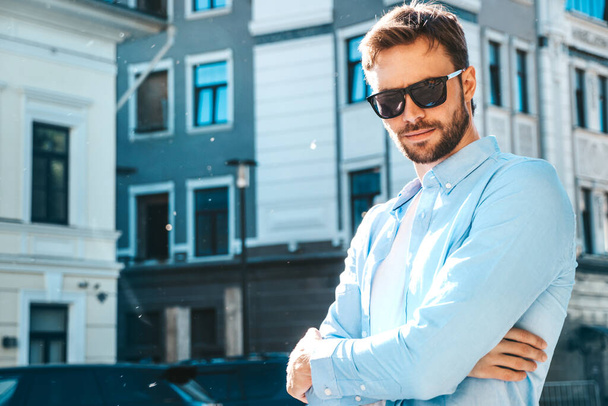 Handsome confident stylish hipster lambersexual model.Modern man dressed in blue shirt. Fashion male posing near skyscraper on the street background in sunglasses. Outdoors at sunset.Crossed arms - Fotoğraf, Görsel