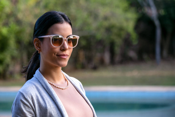 A beautiful brunette woman at the pool wears a dark glasses with a beige lens, a pearl necklace and a neutral-colored female blazer. Social Commitment. Model. Golden hour  - Photo, Image