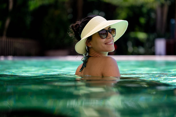 Una hermosa morena disfruta de un día soleado descansando en una piscina de agua caliente con sombrero de paja y gafas oscuras. Láser. Vida sana. Mujer brasileña. Relajación total. Sonríe. - Foto, Imagen