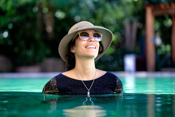 Une belle brune portant un bikini en dentelle noire assorti à un chapeau de paille et des lunettes de soleil pose à l'intérieur d'une piscine. La mode. Une Brésilienne. Loisirs. une vie saine. Bonne journée.. - Photo, image