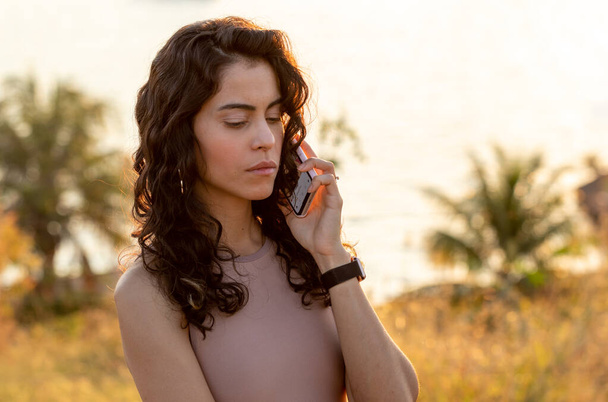 A brunete woman with curly hair at sunset worried talking on the phone at the edge of a lake. Sunny day. Golden hour. Lifestyle. - Foto, Bild