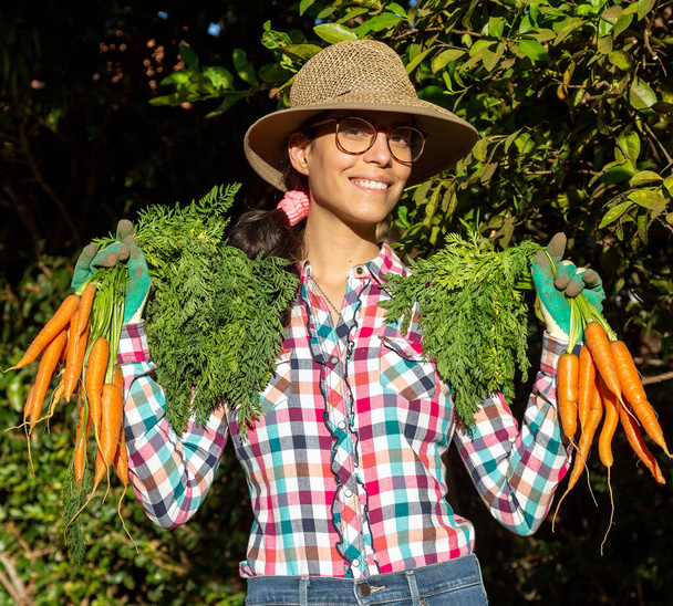 Una morena feliz con las zanahorias que ha cosechado de su jardín orgánico. Lleva gafas graduadas, sombrero de paja y camisa a cuadros. Comida vegana. Vida sana. Estilo de vida. Hora dorada. - Foto, Imagen