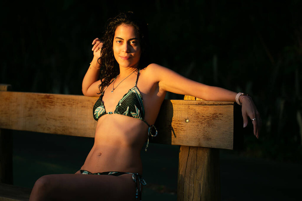 A wonderful brunette woman sitting on a bench next to the pool poses for a photograph at dusk. Golden hour. Brazilian Girl. Leisure. healthy life - Foto, immagini