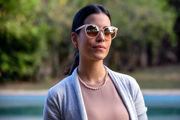 At the pool a beautiful brunette woman wears a dark glasses with a beige lens, a pearl necklace and a neutral-colored female blazer. Social Commitment. Model. Golden hour  - Foto, immagini