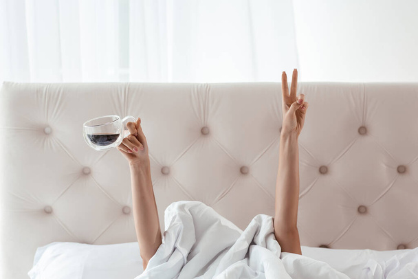 Mão feminina mostrando o sinal engraçado e segurando copo atrás cobertor na cama, conceito da manhã de café. duas mãos de mulher saindo do cobertor. - Foto, Imagem