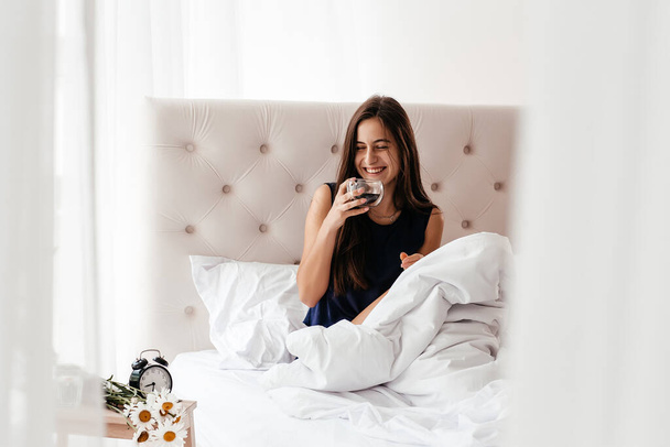 Image of attractive young woman wearing lingerie smiling and drinking coffee in her hands on bed. Coffee morning concept. - Φωτογραφία, εικόνα