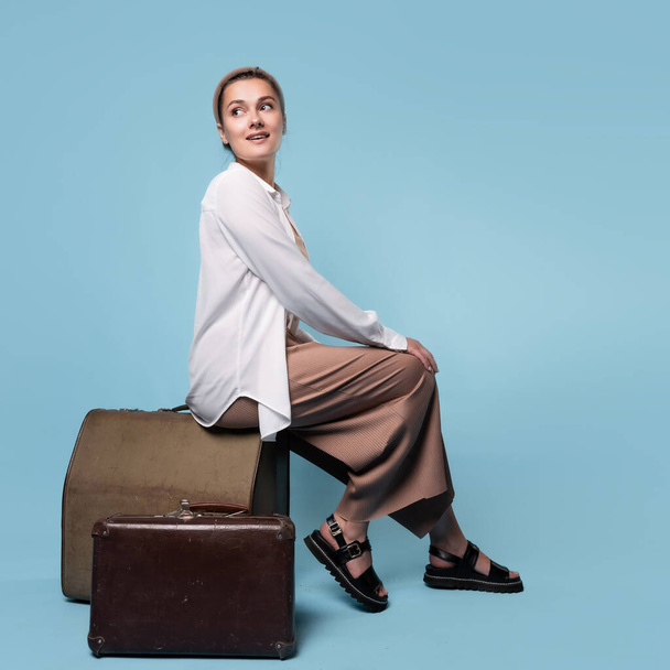 happy young woman in summer pants and a white shirt is sitting on vintage leather suitcases - Foto, immagini