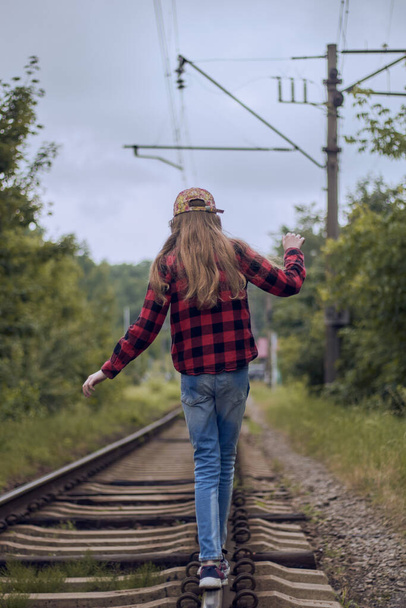 Menina nos trilhos ferroviários. Menina no caminho de ferro em jeans, boné e camisa em uma cela. Vista traseira.  - Foto, Imagem
