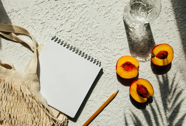 Summer flat lay on a concrete background. Notepad, peaches and an eco-friendly mesh shopping bag. Shadow and sunshine, sun. Summer travel fashion concept. - Фото, изображение