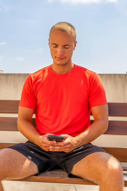 Καυκάσιος νεαρός κάθεται σε ένα παγκάκι και χρησιμοποιεί τηλέφωνο με φόντο τον τοίχο. Τεχνολογική έννοια - Φωτογραφία, εικόνα