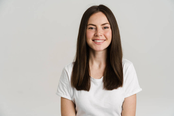 Close up portrait of a smiling young white woman with long brown hair wearing white t-shirt standing over gray background looking at camera - Foto, Bild