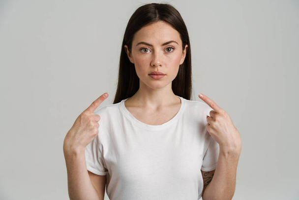 Young brunette woman looking at camera while pointing fingers at herself isolated over white background - Foto, Bild