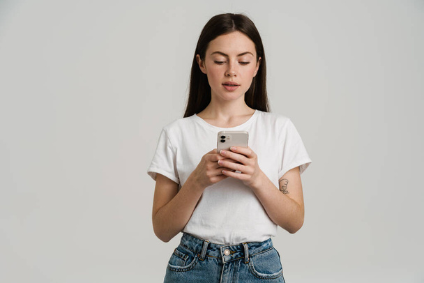 Young brunette woman in t-shirt using mobile phone isolated over white background - Fotoğraf, Görsel