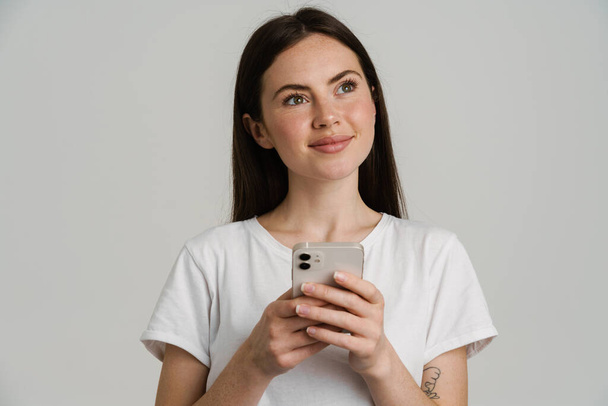 Young brunette woman in t-shirt smiling while using mobile phone isolated over white background - 写真・画像