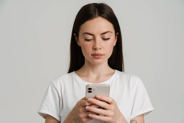 Young brunette woman in t-shirt using mobile phone isolated over white background - Zdjęcie, obraz