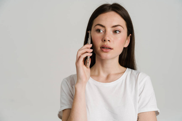 Close up portrait of a young white woman with long brown hair wearing white t-shirt standing over gray background talking on mobile phone looking aside - Photo, image