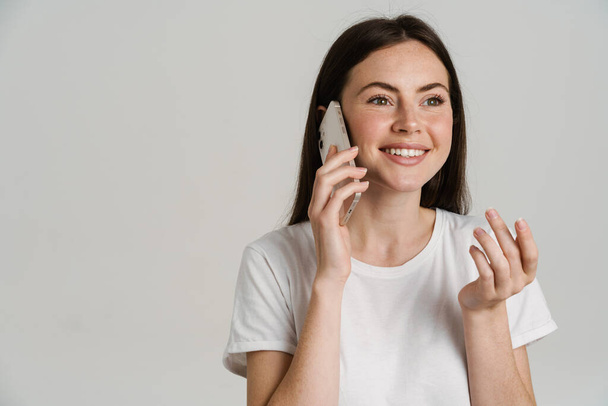Close up portrait of a smiling young white woman with long brown hair wearing white t-shirt standing over gray background talking on mobile phone - Foto, imagen