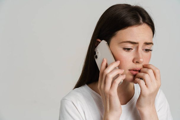 Close up portrait of an upset young white woman with long brown hair wearing white t-shirt standing over gray background talking on mobile phone bad news - Fotoğraf, Görsel