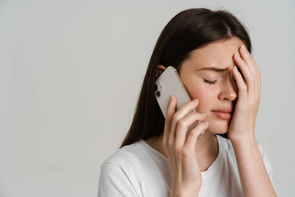 Close up portrait of an upset young white woman with long brown hair wearing white t-shirt standing over gray background talking on mobile phone bad news - 写真・画像
