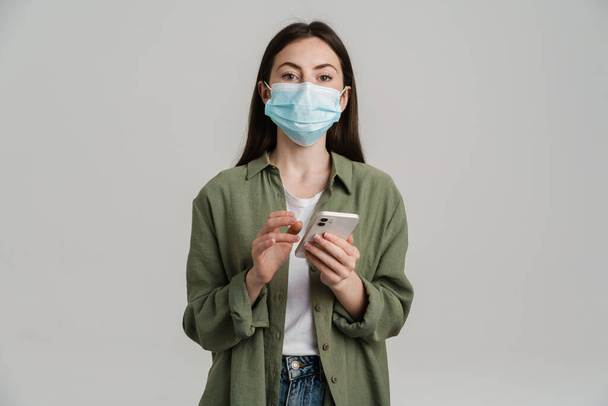 Positive young white woman wearing protective medical mask hoding mobile phone standing over gray wall - Foto, Imagen