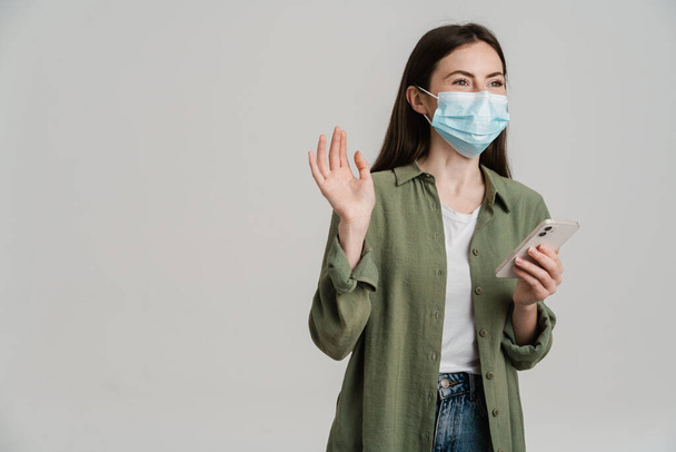 Positive young white woman wearing protective medical mask hoding mobile phone standing over gray wall waving hand - Fotó, kép