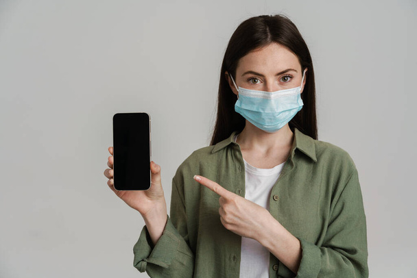 Positive young white woman wearing protective medical mask hoding mobile phone standing over gray wall showing empty screen mobile phone - Fotó, kép