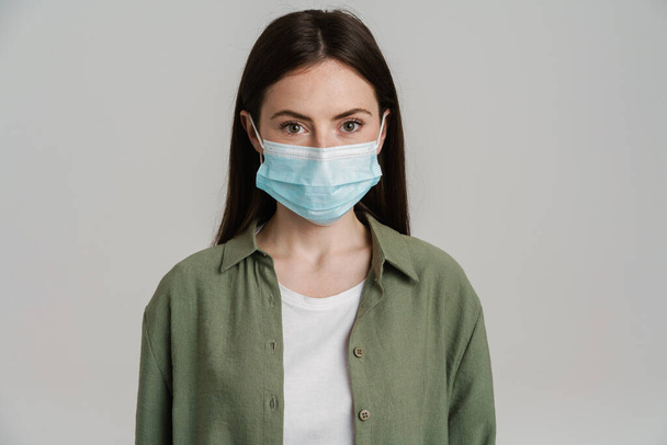 Portrait of a young white casual woman with long brown hair standing wearing protective medical mask over gray wall - Foto, afbeelding