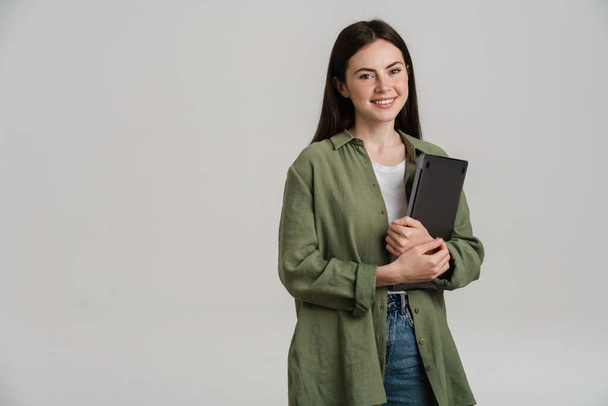 Happy young white casual woman standing holding laptop computer over gray wall background - 写真・画像