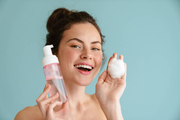 Half-naked brunette woman smiling while showing cleaning foam isolated over blue background - Photo, Image