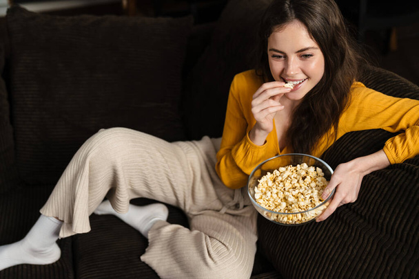 Smiling young brunette woman eating popcorn while sitting on a couch at home - Фото, зображення