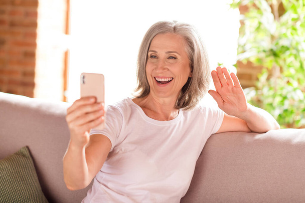 Photo of cute pretty lady pensioner wear white t-shirt sitting sofa smiling talking modern gadget waving arm indoors house flat - Foto, Imagem