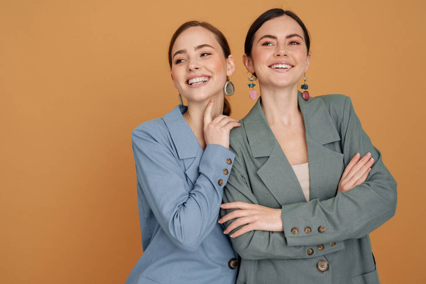 Two smiling elegant women wearing smart jackets posing standing close together isolated over beige background - Zdjęcie, obraz