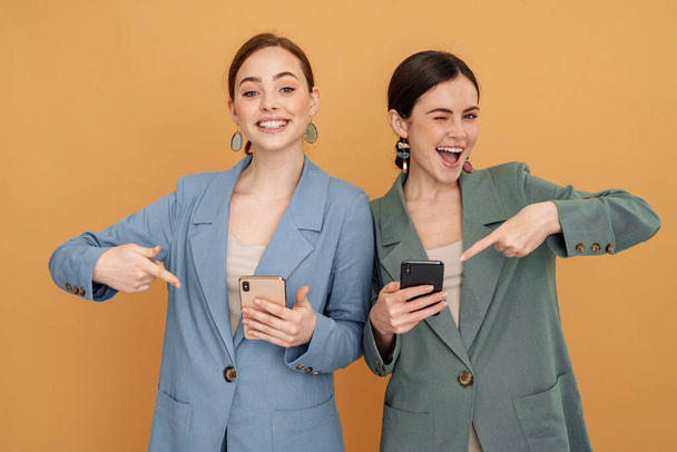 Young two women using and pointing finger at mobile phones isolated over yellow background - Foto, imagen